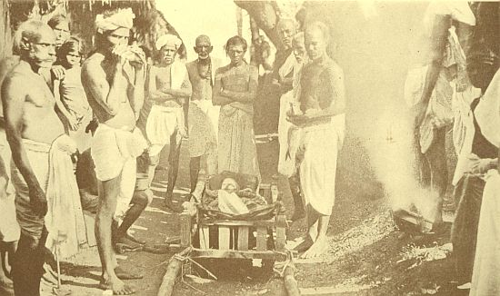 A photo rarely possible. The dead woman lies in her bier; the white on her eyes and brow is the mark of Siva's ashes. Some of the mourners are so marked, as they are all Saivites. The fire is lighted from the pot of fire to the right. Just before it is lighted, the chief mourner takes a vessel of water, pierces a hole in it, walks round the dead, letting the water trickle out, pierces another hole and repeats the walk. After the third piercing and walk, he throws the pot backwards over his shoulder, and as it smashes the water all splashes out. This is to refresh the spirit if it should be thirsty while its body is being burned.