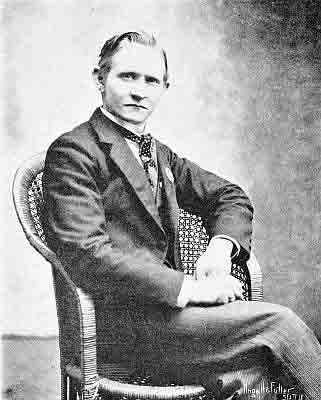 Cravat; dark jacket and vest with narrow pinstripped trousers; seated in cane and bamboo chair.