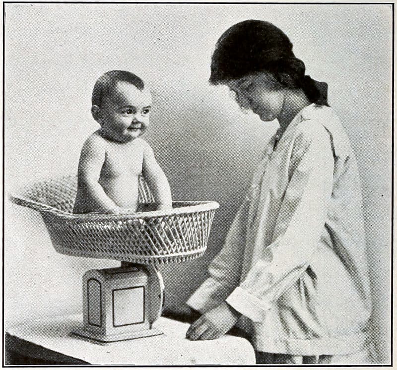 A baby in a basket on a scale that is being read by a woman